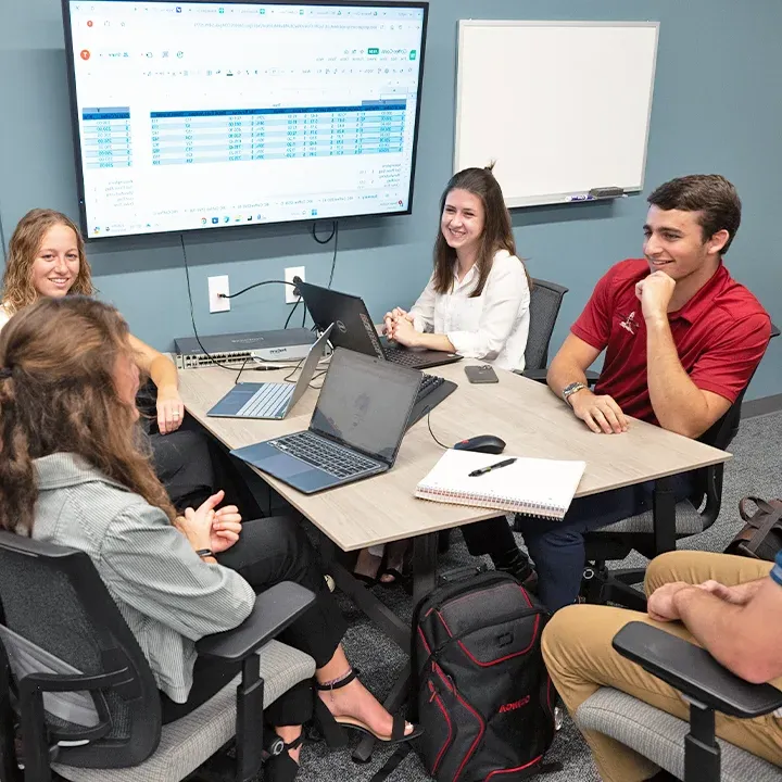 Small group of students gathered at table with laptops and notebooks.