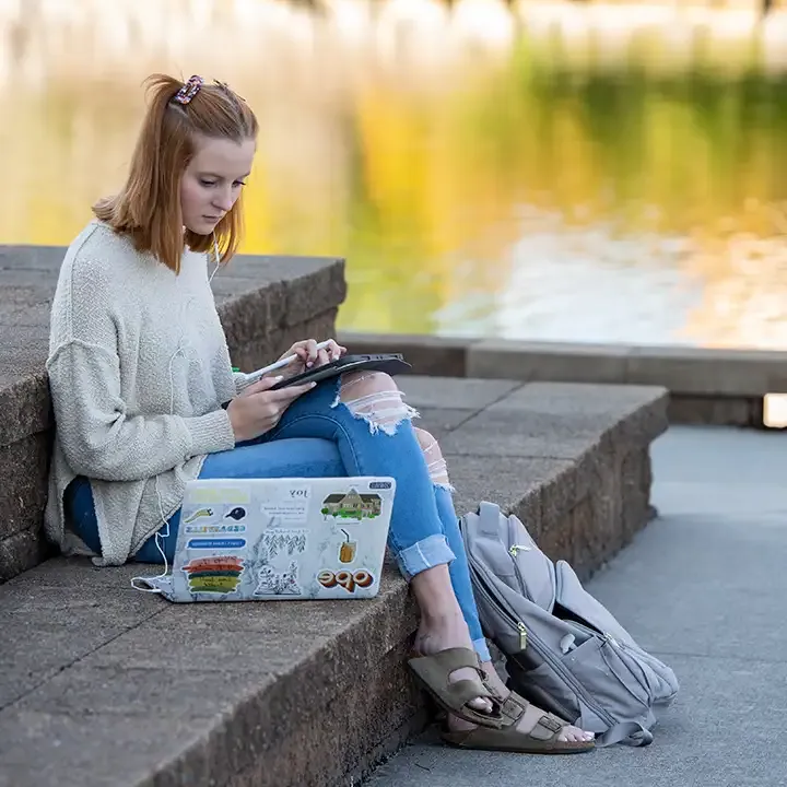 Woman sitting on outside steps by a pond working on laptop.